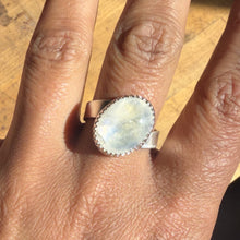 Load image into Gallery viewer, A hand displays the Rainbow Moonstone Twisted Cigar Sterling Silver Ring (Size 9.5), featuring a large oval rainbow moonstone set in a decorative bezel. Shown in bright natural light on a wooden surface, this handmade ring elegantly highlights shimmering hues as it catches the sunlight.