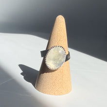 Load image into Gallery viewer, The Rainbow Moonstone Twisted Cigar Sterling Silver Ring (Size 9.5) is a handmade piece featuring a large oval rainbow moonstone in a decorative bezel, shown on a light wooden cone with a white and gray backdrop.