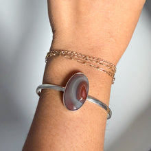 Load image into Gallery viewer, A wrist adorned with the Botswana Agate Cuff in size 5.5, featuring an oval brown and white gemstone set in sterling silver, paired with delicate gold chain bracelets for an elegant display of handmade jewelry.
