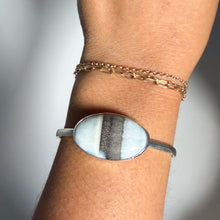Load image into Gallery viewer, A wrist adorned with two gold chain bracelets and the Blue Owyhee Opal Cuff in size 5, featuring a sterling silver band set with an oval stone displaying a gray and white striped pattern.