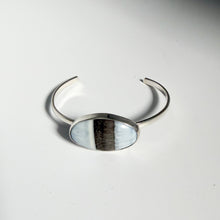 Load image into Gallery viewer, Introducing the Blue Owyhee Opal Cuff | Size 5: a striking sterling silver cuff bracelet showcasing a central oval stone inspired by Blue Owyhee Opal, its surface accented with a vertical black stripe bordered by delicate gradients of white and pale blue.