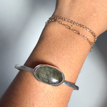 Load image into Gallery viewer, A wrist adorned with the Labradorite Cuff in size 6, featuring a striking oval greenish gemstone set in sterling silver, alongside two delicate gold chain bracelets, showcases elegant handmade jewelry.