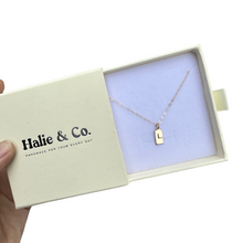 Load image into Gallery viewer, A hand holds an open cream-colored Halie & Co. jewelry box containing the Alphabet Initial Rectangle Necklace, which features a gold filled rectangular pendant engraved with the letter L.